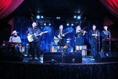 Six musicians of Roselit Bone prepare for their performance during a pre-show soundcheck at the intimate Dante's venue in Portland, Oregon. The band assembles on stage beneath dramatic blue and red theatrical lighting, with instruments including electric guitars, violin, and keyboards creating an atmospheric tableau. Crimson curtains frame the scene while professional stage lights pierce through the venue's characteristic darkness, capturing the focused energy of rehearsal at this legendary Portland music club.