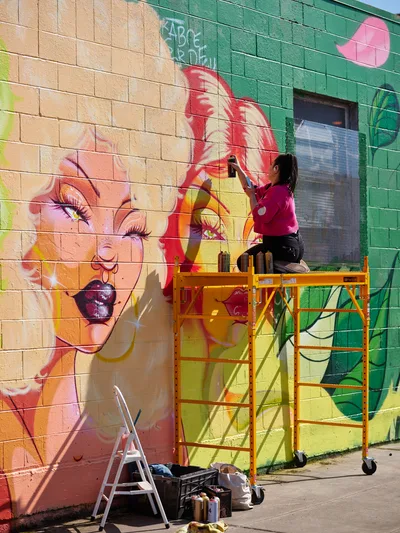 Multidisciplinary artist Vilatose works from an orange scaffolding platform, adding vibrant details to a large-scale mural on Northeast Alberta Street in Portland's Alberta Arts District. The colorful artwork features a portrait of a woman with flowing red hair and dramatic makeup against a backdrop of warm peach and emerald green blocks. Afternoon sunlight casts sharp shadows across the concrete sidewalk, where paint supplies and a step ladder await below the industrial mobile scaffold.