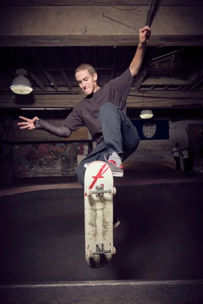 A young skateboarder launches into motion beneath the weathered wooden beams of Commonwealth Skateboarding's subterranean skate space in Portland's Buckman neighborhood. Illuminated by industrial pendant lights, the athlete grips his white skateboard marked with a distinctive red 'F' logo while suspended mid-trick against the basement's rustic architecture. The raw concrete ceiling and exposed timber framework create a gritty urban sanctuary where street skating culture thrives in the shadows.