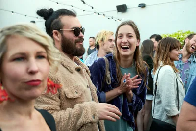 Under the soft glow of string lights at Portland's White Owl Social Club, a diverse crowd of young adults celebrates at Your Sunday Best's season opener. A bearded man in sunglasses and sherpa jacket shares an animated moment with a laughing brunette woman in navy velvet, while a blonde woman in a textured coat occupies the foreground. The outdoor patio buzzes with the energy of summer socializing, capturing the spontaneous joy of Portland's vibrant day party scene.