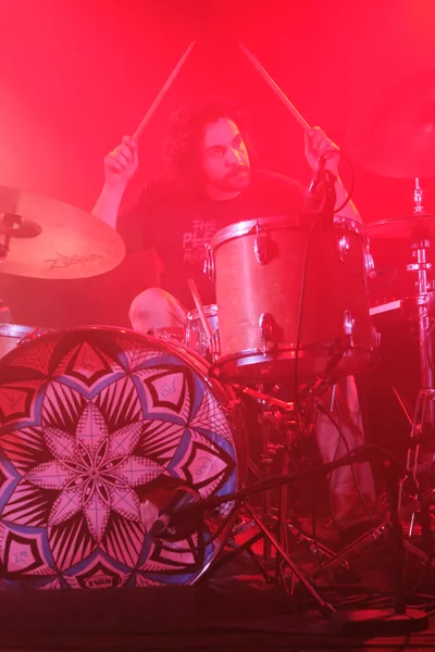 A drummer from Sun Blood Stories emerges through dense red stage lighting at Dante's nightclub in Portland's downtown district. The performer, captured mid-beat with drumsticks raised, appears almost spectral behind the thick crimson fog that transforms the intimate venue into a fever dream. An ornate mandala-patterned drum head anchors the foreground while the atmospheric haze swallows the drum kit's chrome hardware, creating an otherworldly concert experience that defines Portland's underground music scene.
