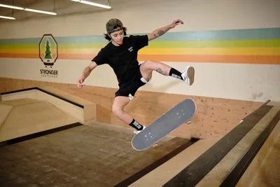 Professional skateboarder Christiana Means executes a precise aerial maneuver above the wooden bowl at Stronger Skatepark in Milwaukie, Oregon. Suspended mid-flight with arms extended for balance, she demonstrates complete control as her skateboard hovers beneath her. The indoor facility's warm lighting illuminates the rainbow-striped mural backdrop, creating a dynamic contrast against the natural wood construction of the skate bowl.