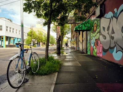A blue bicycle rests against weathered concrete beside vibrant murals adorning the former Oregon Theater on Southeast Division Street in Portland's Richmond neighborhood. The street art transforms the aging storefront with bold greens, pinks, and blues, while leafy trees filter soft daylight across the quiet sidewalk. The juxtaposition of urban decay and artistic renewal captures the evolving character of this historically industrial Portland corridor.