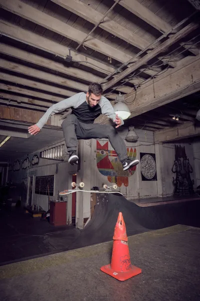 A skateboarder launches into the air above a bright orange traffic cone in the dimly lit Commonwealth Skateboarding in Portland's Buckman neighborhood. The raw industrial space features exposed wooden ceiling beams, graffiti-covered walls, and makeshift ramps, creating an authentic underground skate culture atmosphere. Moody lighting from overhead fixtures casts dramatic shadows across the concrete floor, while colorful street art and skateboard graphics provide vibrant contrast against the weathered interior.
