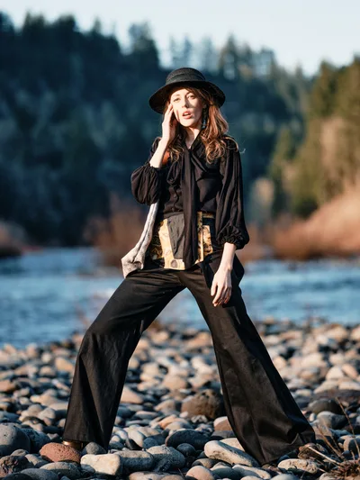 A woman in flowing black attire and wide-brimmed hat stands contemplatively on the rocky shoreline of the Columbia River along Multnomah County's Floodplain Trail. The golden hour light illuminates her thoughtful pose as she raises one hand to her temple, her wide-leg trousers and billowing blouse creating dramatic silhouettes against the water. The background reveals the characteristic forested hills of the Columbia River Gorge, with smooth river stones scattered beneath her feet on the exposed riverbed.