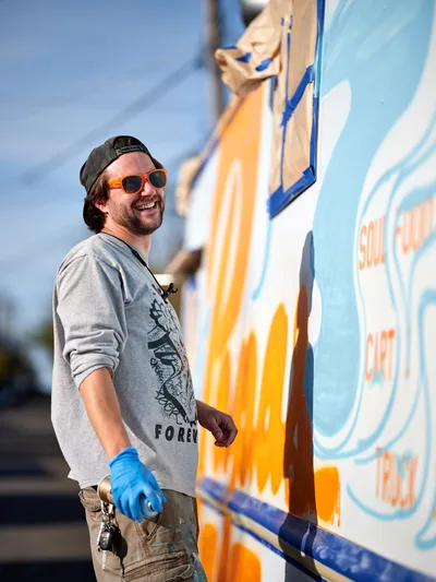 A bearded muralist in orange sunglasses and a backwards cap grins while working on a colorful food truck exterior in Portland's King neighborhood. Wearing blue protective gloves and a gray graphic sweatshirt, he stands before his vivid orange and blue artwork that transforms the vehicle's surface. The bright afternoon sunlight illuminates both the artist and his dynamic street art, capturing the creative energy of Portland's thriving food truck culture.