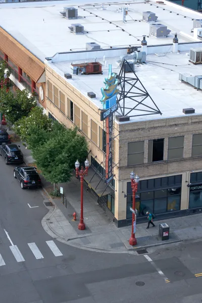 An elevated perspective captures the intersection where traditional meets contemporary in Portland's Chinatown district. The geometric precision of white crosswalk stripes guides the eye through a scene where vintage brick architecture houses modern commerce, while HVAC units dot the pristine white rooftop like mechanical sentinels. Red street lamps stand as colorful punctuation marks against the muted palette of concrete and brick, their warm hue echoing the neighborhood's cultural heritage.