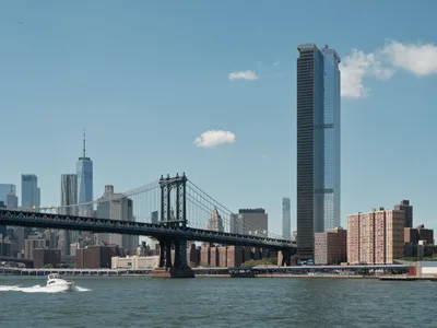 The iconic Manhattan Bridge stretches across the East River in elegant suspension, its steel towers and cables creating geometric lines against a pale blue afternoon sky dotted with cotton-white clouds. One World Trade Center's gleaming spire punctuates the left side of Manhattan's financial district, while a striking dark glass tower dominates the right portion of the frame. A white pleasure boat cuts through the choppy river waters in the foreground, leaving a gentle wake as it passes beneath this architectural symphony of old and new New York.