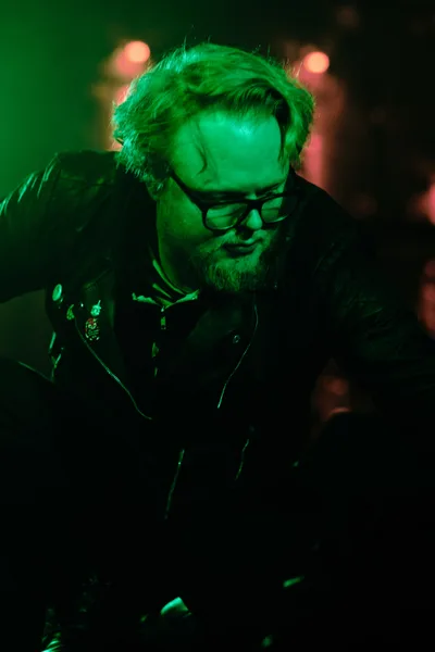 Electronic musician Eli Savage of Ellipsism performs under intense emerald stage lighting at Dante's in Portland's downtown district. The atmospheric green wash creates a cyberpunk aesthetic, casting the bearded artist in an otherworldly glow as he concentrates over his equipment. Warm bokeh lights pierce through the darkness behind him, while his leather jacket and glasses catch the saturated color, creating a moody portrait of underground electronic music culture.