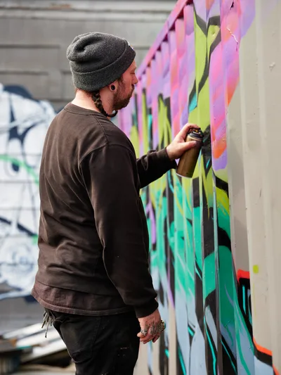 A bearded artist in a charcoal beanie and brown hoodie concentrates intently as he applies spray paint to a colorful mural on a concrete wall in Northeast Portland, Oregon. The artwork features flowing organic shapes in brilliant pinks, greens, and yellows that contrast sharply against the gray urban surface. Soft natural light illuminates the scene, capturing the focused energy of creative expression in an industrial setting. Additional graffiti work is visible in the background, establishing this as part of Northeast Portland's thriving street art scene.