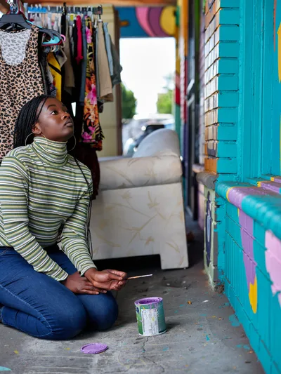 Naomi Likayi sits contemplatively on the concrete sidewalk beside her vibrant turquoise and purple mural in Northeast Portland, preparing to sign her completed artwork. Dressed in a striped turtleneck and denim, she holds a paintbrush while a purple paint can rests nearby, capturing a moment of artistic completion in this colorful Alberta Arts District storefront setting. The scene frames her against hanging garments and the sun-drenched street beyond, embodying the creative spirit that defines this neighborhood's cultural landscape.