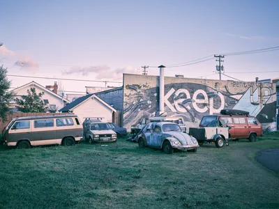 A collection of aging and worn vehicles, including two Volkswagen Vanagons, a rusted VW Beetle, and a red van with a roof rack, are parked on a grassy lot in Portland's Alberta Arts District. Behind them, a large weathered building displays a partial mural reading "Keep...Up," part of the "Keep Your Chin Up" street art piece, featuring painted feathers and organic forms in black, white, and blue. Residential homes, utility poles, and a soft purple-hued evening sky are visible in the background.