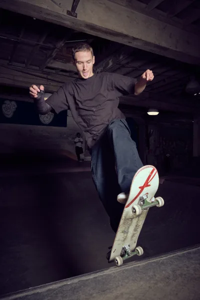 A skateboarder executes a fluid maneuver in the shadowy concrete bowl at Commonwealth Skateboarding in Portland's Buckman neighborhood. Dressed in a dark sweatshirt and loose jeans, the rider navigates the curved transition with focused intensity, his weathered deck bearing a distinctive red lightning bolt graphic. The underground skate park's exposed concrete beams and industrial lighting create a gritty urban cathedral for this moment of athletic grace.