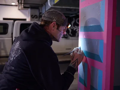Portland artist and bike activist Rabbitt meticulously applies paint to a vibrant geometric mural in the dimly lit parking garage of the Clay Creative building in the Hosford-Abernethy neighborhood. Wearing a camo cap and dark hoodie, the tattooed artist works with focused precision on bold pink, blue, and purple angular forms that transform the industrial concrete space. The fluorescent lighting creates dramatic shadows as he brings color and artistry to the urban underground environment.
