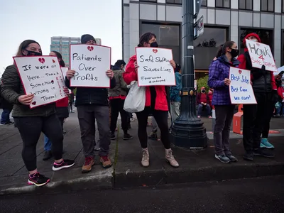 Healthcare workers gather on a grey Portland morning outside the Kaiser Permanente Tower in the Lloyd District, their handmade protest signs creating stark white rectangles against the building's glass facade. Masked nurses in winter jackets hold placards declaring "Patients Over Profits" and "Safe Staffing Saves Lives," their determination evident despite the overcast sky. The urban sidewalk scene captures the tension between corporate healthcare and frontline workers, with the tower's reflective windows looming behind the small but resolute group of demonstrators.