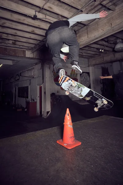 A skateboarder defies gravity in mid-air, soaring over a bright orange traffic cone inside the weathered confines of Commonwealth Skateboarding in Portland's Buckman neighborhood. The exposed wooden ceiling beams and graffitied walls create a gritty urban backdrop as the athlete executes a precise aerial maneuver, skateboard clutched firmly beneath gray-clad legs. Industrial lighting cuts through the basement-like atmosphere of this Southeast Portland skate haven, capturing the raw energy and creative spirit that defines the local skateboarding scene.