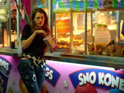 A young woman with auburn hair reaches into a glowing popcorn cart at the Clark County Fair in Vancouver, Washington. The warm golden light from the concession stand contrasts with the cool purple and blue neon of carnival rides spinning in the background. Her striped shirt and distressed jeans capture the casual energy of fairgoers, while the scene pulses with the electric atmosphere of late-night carnival revelry.