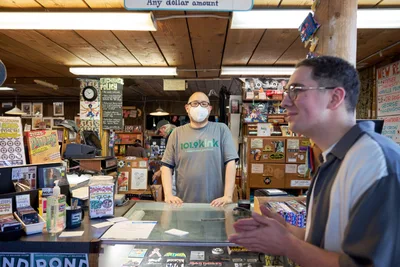 Inside Music Millennium on Portland's East Burnside during Record Store Day 2022, a masked store employee in a gray t-shirt stands behind the glass counter while engaging with a customer. The intimate record store atmosphere is illuminated by warm overhead lighting, revealing walls densely packed with vinyl records, vintage posters, and music memorabilia. The wooden beam ceiling and cluttered shelving create an authentic, lived-in feeling that captures the essence of Portland's independent music culture during this annual celebration.