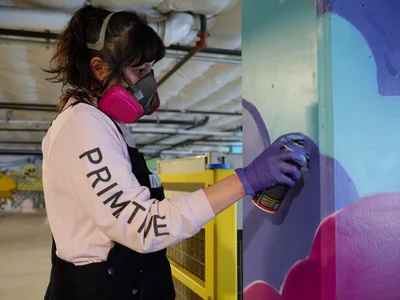A focused muralist wearing protective gear and purple gloves carefully applies spray paint to a vibrant wall inside Clay Creative's parking garage in Southeast Portland. The artist, dressed in a pink 'PRIMETIME' sweatshirt and respirator mask, works methodically on flowing abstract forms in blues and purples. Industrial ceiling fixtures cast even light across the underground creative space, while yellow structural elements provide stark contrast to the emerging artwork.