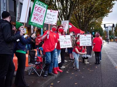 Healthcare workers in crimson masks and shirts gather on Northeast Multnomah Street outside Kaiser Permanente Tower, their handmade signs demanding fair wages and union recognition piercing the overcast Portland evening. The AFSCME banners and personal placards create a sea of red against the urban backdrop, while folding chairs and determined faces signal a prolonged demonstration. Autumn leaves scatter across wet pavement as protesters maintain their vigil beneath the towering medical complex, their collective voice rising in the Lloyd District's fading light.