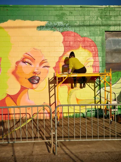Multidisciplinary artist Vilatose works from a yellow scaffolding platform, adding details to Jardín Cubana, a vibrant mural featuring three female figures on the brick wall at Donny Vegas in Portland's Alberta Arts District. The mural bursts with warm yellows, coral pinks, and emerald greens against the industrial backdrop of metal barriers and urban streetscape. Late afternoon light bathes the scene as the artist, dressed in a mustard yellow sweatshirt, carefully refines the figurative composition with concentrated precision.