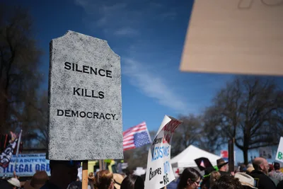 A weathered granite tombstone bearing the stark inscription 'SILENCE KILLS DEMOCRACY' towers above a sea of protesters gathered in Portland's Chinatown district. The mock gravestone, held aloft against a crisp blue sky dotted with white clouds, serves as the focal point of the March 2026 No Kings demonstration. American flags flutter among the crowd while various protest signs create a mosaic of dissent, including visible text about resistance and legal obligations.