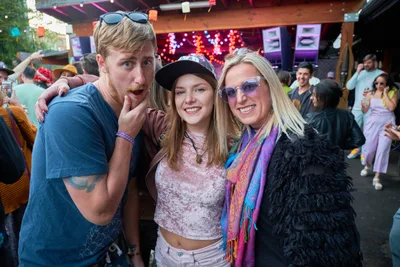 Three festival-goers embrace in a moment of pure joy at White Owl Social Club's Your Sunday Best day party in Portland, Oregon. The trio - a bearded man in blue flannel making a playful gesture, a young woman in bohemian attire with a black cap, and a blonde woman draped in a vibrant shawl and black feathers - radiate infectious energy against the backdrop of string lights and warm wooden architecture. The outdoor venue pulses with summer festival atmosphere as other revelers dance and socialize in the golden afternoon light.