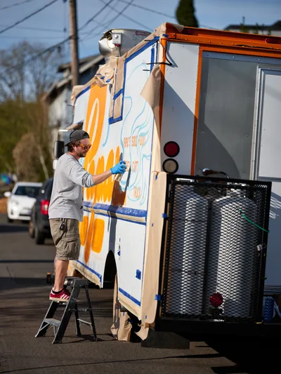 A bearded muralist in casual attire stands on a stepladder, spray-painting vibrant orange and blue designs onto the side of a white food truck parked along Northeast 9th Avenue in Portland's King neighborhood. The afternoon light illuminates his concentrated work as protective plastic sheeting flutters around the vehicle's windows. Power lines and residential buildings create an urban backdrop typical of Portland's eclectic food truck culture.