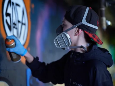 A young artist wearing a respirator mask and baseball cap carefully applies spray paint to a vibrant mural in the industrial confines of Clay Creative's parking garage in Portland's Hosford-Abernethy neighborhood. The atmospheric lighting catches swirls of aerosol mist as colorful graffiti blooms across concrete walls, transforming the utilitarian space into an underground canvas. Blue-gloved hands guide the spray can with practiced precision while neon hues pierce through the shadowy garage interior.