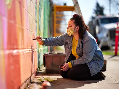 Multidisciplinary artist Villatose sits cross-legged on the sidewalk, brush in hand, adding intricate details to her 'Jardín Cubana' mural featuring three female figures along Alberta Street in Portland's arts district. Golden afternoon light bathes the scene as she works methodically on the colorful wall, her floral face mask and mustard sweater creating a striking contrast against the weathered denim jacket. The composition captures the intimate concentration of street art creation, with urban elements softly blurred in the background.