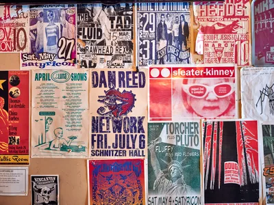 A densely layered collage of weathered concert posters and flyers creates a visual time capsule of Portland's indie music scene on the wall of Foster Burger. The overlapping promotional materials showcase bands like Dan Reed Network, Sleater-Kinney, and various underground acts, their faded colors and torn edges telling stories of late-night shows at venues like La Luna and Satyricon. The chaotic arrangement pulses with the raw energy of DIY concert promotion, each poster a artifact of the city's vibrant cultural underground.