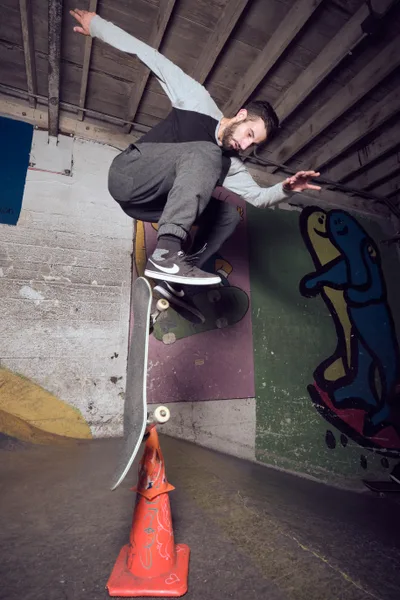 A bearded skateboarder in gray sweatpants and white hoodie executes a trick over a red traffic cone in the gritty underground confines of Commonwealth Skateboarding in Portland's Buckman neighborhood. The raw concrete walls are adorned with colorful graffiti art, while weathered wooden ceiling beams cast dramatic shadows across the subterranean skate sanctuary. His focused expression and extended arms capture the precise balance required as he navigates the urban obstacle course beneath the city streets.