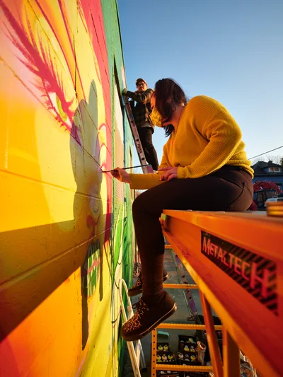 Multidisciplinary artist Vilatose works on the dynamic mural Jardín Cubana from an orange Metaltec scissor lift on Alberta Street in Portland's arts district. The late afternoon light bathes the scene in warm golden tones as the artist, wearing a mustard yellow sweater, carefully applies paint to the bold composition depicting three female figures spanning bright yellows, coral pinks, and lime greens. A second collaborator assists from above while the Alberta Arts District's creative energy pulses through this moment of urban art creation.