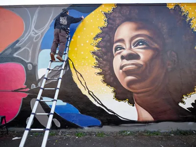 Street artist Travis Fields, known as Campographic, meticulously works from an aluminum ladder to paint a vibrant portrait of poet Amanda Gorman on a concrete wall in Portland's Alberta district. The mural captures Gorman's likeness with remarkable detail, featuring her natural hair crowned with a golden halo against a backdrop of swirling colors and abstract elements. The scene unfolds on Northeast Killingsworth Street, where the artist's concentrated effort transforms the urban canvas into a celebration of literary achievement.