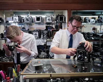 In the intimate confines of Blue Moon Camera & Machine, Portland's legendary camera haven, Mike Knight and a colleague work with reverent concentration behind a gleaming glass counter. The warm tungsten light illuminates their careful examination of vintage equipment while towering displays of classic cameras—from twin-lens reflex models to large format view cameras—create a museum-like backdrop. The scene captures the quiet dedication of analog photography's guardians, their focused gestures speaking to decades of expertise amid this treasure trove of mechanical precision.