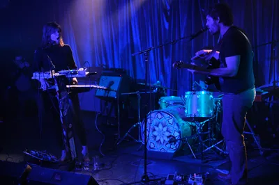 Boise experimental duo Sun Blood Stories performs in the intimate confines of Dante's nightclub in downtown Portland during Rx Fest. Bathed in ethereal blue and purple stage lighting, a keyboardist and drummer create an atmospheric soundscape against flowing curtain backdrops. The moody illumination transforms the underground venue into a dreamlike performance space, with the duo's silhouettes emerging from the saturated concert lighting as they craft their modern-day operatic compositions.
