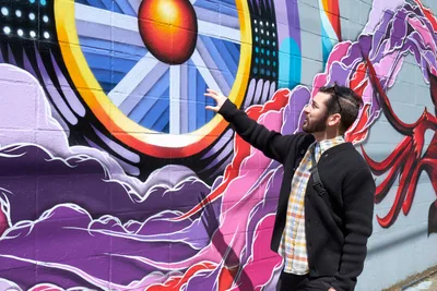 Tomás Valladares of the Portland Street Art Alliance reaches toward a vibrant mural during an interpretive tour in Portland's Central Eastside district. The dramatic wall art features swirling purple and pink forms surrounding a luminous orange orb, creating a dynamic cosmic landscape against the concrete surface. Valladares, dressed in a dark sweater and plaid shirt with a messenger bag, gestures enthusiastically as he shares the story behind the artwork. The scene captures the intersection of community education and urban artistry on Southeast 11th Avenue.
