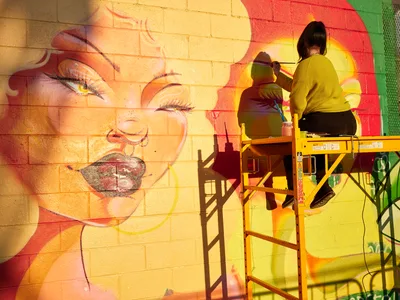 Multidisciplinary artist Villatose works from a yellow scissor lift, painting details on Jardín Cubana, a vibrant mural featuring three female figures in warm yellows and coral tones. The artist, wearing a mustard yellow sweater, carefully applies brushstrokes to the large-scale portrait on the brick wall of a building in Portland's Alberta Arts District. Sharp afternoon shadows create dramatic geometric patterns across the colorful street art, emphasizing the contrast between the industrial lift and the flowing, organic forms of the emerging artwork.