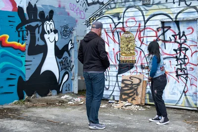 Two visitors stand before a vibrant mural wall in Portland's Central Eastside during a guided street art tour. The weathered concrete surface displays layers of graffiti including a whimsical cartoon character, ornate lettering, and colorful geometric forms. The tour guide and participant examine the urban artwork amid scattered debris on the industrial pavement, capturing the raw authenticity of this evolving neighborhood's creative expression.