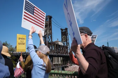 Protesters gather beneath Portland's Steel Bridge on a brilliant spring day, raising American flags and yellow signs declaring "Democracy Not Autocracy" against the industrial lattework of the historic lift bridge. The scene captures citizens exercising their First Amendment rights with the Willamette River waterfront as backdrop, while the bridge's towering steel framework creates dramatic geometric patterns against the azure sky. Demonstrators in casual attire hold their signs high, their message echoing across the water toward downtown Portland's skyline visible in the distance.