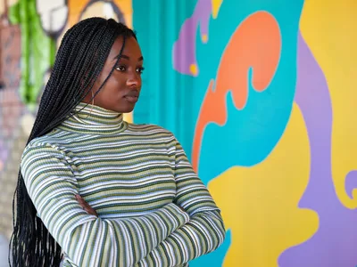 Naomi Likayi stands confidently with crossed arms before her vibrant street mural in Portland's Alberta Arts District. Her long box braids cascade over a striped mock-neck sweater as she gazes thoughtfully over her shoulder. The explosive backdrop features bold organic shapes in turquoise, coral orange, sunny yellow, and purple, creating a dynamic contrast against the urban setting. Soft natural light illuminates her face, capturing a moment of artistic accomplishment and quiet pride.