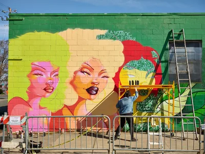Multidisciplinary artist Vilatose applies vibrant pigments to 'Jardín Cubana,' a larger-than-life portrait featuring three female figures on Northeast Alberta Street, transforming a green cinderblock wall into a kaleidoscope of feminine faces rendered in electric pinks, corals, and golden yellows. Working from scaffolding beneath Portland's crystalline winter sky, the artist adds delicate details to expressive eyes and glossy lips while construction barriers frame the creative process. The emerging mural radiates pop art sensibilities against the industrial backdrop of the Alberta Arts District.