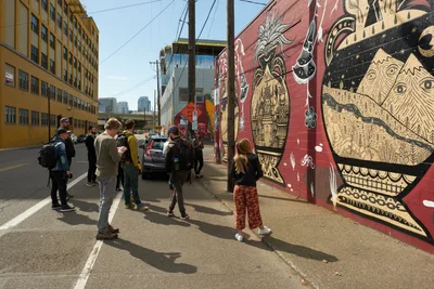 A diverse group of street art enthusiasts gathers on Southeast Stark Street in Portland's Central Eastside, their shadows stretching across weathered asphalt as they contemplate an intricate mural. The towering artwork features mystical creatures and celestial motifs in warm terracotta and cream tones, while the industrial streetscape of yellow brick buildings and power lines frames this moment of cultural discovery. The afternoon light creates dramatic silhouettes of the tour participants, each absorbed in studying the elaborate wall art that transforms this urban corridor into an open-air gallery.