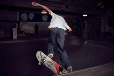 A skateboarder executes a fluid carve through the concrete bowl at Commonwealth Skateboarding in Portland's Buckman neighborhood. The rider, dressed in a crisp white shirt and dark jeans, demonstrates precise board control while navigating the curved transition. Moody tungsten lighting creates dramatic shadows across the weathered concrete surfaces, while fellow skaters observe from the background of this authentic skate sanctuary.