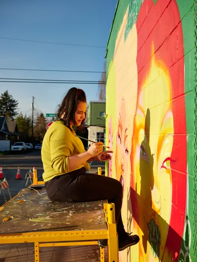Multidisciplinary artist Vilatose sits cross-legged on a yellow scaffold platform, delicately painting details on her mural titled Jardín Cubana that depicts three female figures on the side of a building in Portland's Alberta Arts District. Golden hour light bathes the scene, casting warm shadows and illuminating her yellow cardigan as she works with focused concentration. The mural features flowing bands of red, orange, yellow, and green pigments that seem to dance across the concrete canvas. Behind her, the quiet residential street stretches toward the horizon under a clear blue sky, dotted with power lines and evergreen trees.