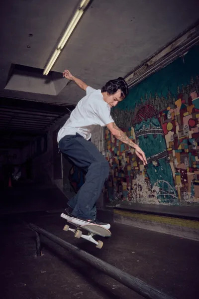 A skateboarder carves through the shadows of Commonwealth Skateboarding's indoor park in Portland's Buckman neighborhood, his white shirt stark against the moody concrete tunnel. Fluorescent lighting cuts harsh angles through the subterranean space while vibrant graffiti murals create a kaleidoscope backdrop of urban art. The rider's fluid motion and focused expression capture the meditative rhythm of street skating in this iconic Pacific Northwest skate sanctuary.