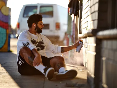 Muralist Heysus sits cross-legged on the sidewalk at Northeast 15th and Killingsworth, spray paint can in hand, bathed in warm golden hour light. The tattooed artist wears earbuds and a graphic tee while working on his latest piece, with colorful graffiti walls and a white van creating an authentic urban backdrop. Long shadows stretch across the concrete as the setting sun illuminates this moment of creative focus in Portland's Alberta Arts District.