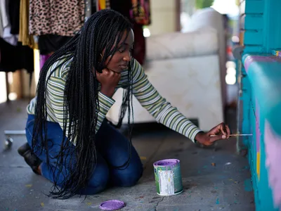 Naomi Likayi sits cross-legged on the concrete sidewalk, delicately applying her signature to a vibrant turquoise mural panel. Her long braids cascade over a striped shirt as afternoon light filters through the urban marketplace atmosphere of Portland's Alberta Arts District. An open paint can with purple pigment sits beside her, evidence of the creative work just completed. The scene captures an intimate moment of artistic completion against the backdrop of Northeast Portland's culturally rich community.