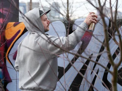 Travis Fields, known as Campographic, concentrates intently on his mural work at the Clay Creative Building in southeast Portland's Hosford-Abernethy neighborhood. Clad in a gray hoodie and snapback cap, the artist grips his spray paint can with focused determination against the concrete wall. Bare winter branches frame the scene in the foreground, while vibrant orange and purple graffiti elements provide a colorful backdrop to this moment of urban artistic creation.