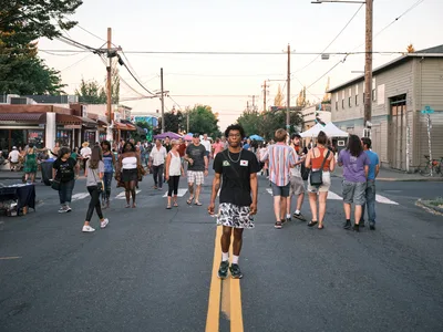 Golden hour light bathes the bustling Last Thursday street fair on Northeast Alberta Street in Portland's Alberta Arts District. A young Black man in glasses and a black t-shirt stands confidently at the center of the closed street, perfectly positioned on the yellow center line, while dozens of festival-goers browse vendor booths and socialize behind him. The scene captures the vibrant community energy of this monthly cultural celebration, with colorful umbrellas, local storefronts, and power lines creating an authentic urban Portland streetscape.